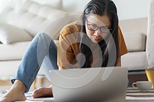 Asian woman wear glasses and casual suite  is working on laptop.Women sitting on the floor with a sofa in the back her in the