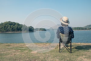 Asian woman wear hat and sitting relax on portable chair nearly lake at National Park.