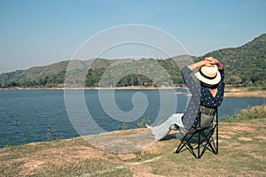 Asian woman wear hat and sitting relax on portable chair nearly lake at National Park.