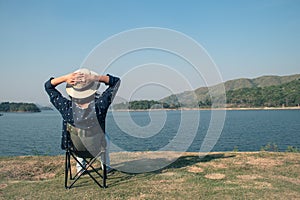 Asian woman wear hat and sitting relax on portable chair nearly lake at National Park.