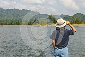 Asian woman wear hat and standing relax on green grass meadow field, she looking forward to lake and mountain.