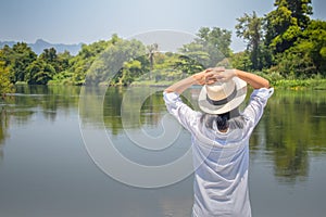 Asian woman wear hat and white shirt with standing on wooden bridge, she looking forward to river with put hers hand on head and s