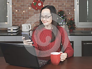 Asian woman wearing eyeglasses ,red knitted sweater sitting with computer laptop in ithe kitchen with Christmas decoration,
