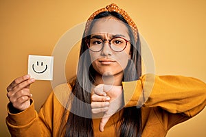 Asian woman wearing glasses holding reminder with smile emoji draw over yellow background with angry face, negative sign showing