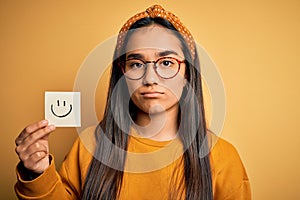Asian woman wearing glasses holding reminder with smile emoji draw over yellow background with a confident expression on smart