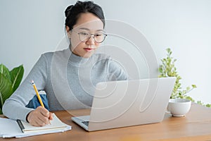 Asian woman wearing glasses is using laptop at home office.Portrait young cute student working on smart technology gadget