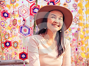 Asian woman wearing hat , sitting among colorful tung flag traditiona north-eastern Thai craft smiling and looking away