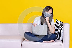 Asian woman wearing medical mask and working from home use laptop computer while sitting on sofa over isolate yellow background
