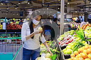 Asian woman wearing protective face mask hold paper shopping bag with fruits and vegetables in supermarket department store. Girl