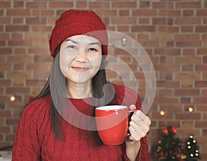 Asian woman wearing red knitted sweater and hat standing in the kitchen decorated with Christmas tree drinking coffee from red mug