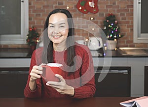Asian woman wearing red knitted sweater sitting in the kitchen decorated with Christmas tree drinking coffee from red mug, smiling