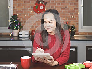 Asian woman wearing red knitted sweater sitting at table with red cup of coffee and gift boxes in the kitchen with Christmas