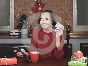 Asian woman wearing red sweater sitting with red cup of coffee and gift boxes in the kitchen with Christmas decoration, holding