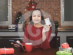 Asian woman wearing red sweater sitting with red cup of coffee and gift boxes in the kitchen with Christmas decoration, holding