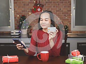 Asian woman wearing red sweater sitting with red cup of coffee and gift boxes in the kitchen with Christmas decoration, holding