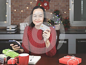 Asian woman wearing red sweater sitting with red cup of coffee, notebook and gift boxes in the kitchen with Christmas