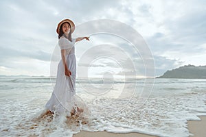 Asian woman wearing stylish hat and clothes pointing at the sky and looking to the ocean and enjoying beautiful sunset on the