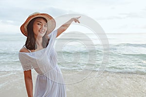 Asian woman wearing stylish hat and clothes pointing at the sky and looking to the ocean and enjoying beautiful sunset on the