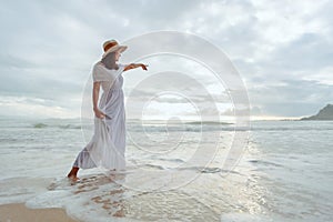 Asian woman wearing stylish hat and clothes pointing at the sky and looking to the ocean and enjoying beautiful sunset on the