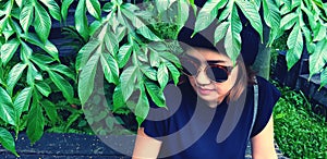 Asian woman wearing sunglass and wool hat sitting on wooden bench under tree with green plant background.