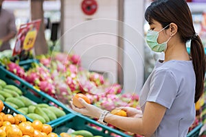 Asian woman wearing surgical face mask and holding orange fruit in supermarket or grocery, protect coronavirus inflection. Hygiene