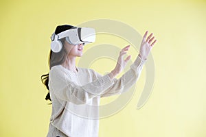 Asian woman wearing virtual reality, VR headset and trying to touch something with hand while stand over isolated yellow