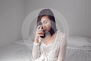 Asian woman in white nightgown drinking water on the bed at bedroom