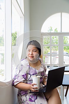An Asian woman in a white patterned dress looking at outside window while working with laptop on a table in a coffee shop