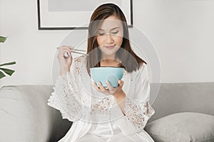 Asian woman in white satin nightgown sitting on a gray sofa and eating food during the night