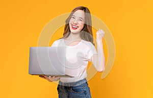 Asian woman in white t-shirt and holding laptop with raises arms and fists clenched with shows strong powerful