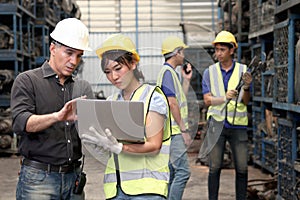 Asian woman worker with safety vest and helmet holds laptop, reports information to her supervisor senior foreman at manufacturing
