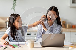 Asian woman working on laptop computer from home while daughter distracting her, taking off mom's glasses