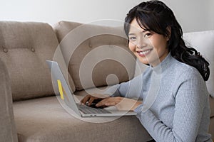 Asian woman is working on a laptop in  living room. She sit on the floor and sofa is office desk. On her face are smile and