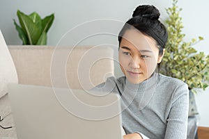 Asian woman is working on a laptop in  living room. She sit on the floor and sofa is office desk. On her face are smile and