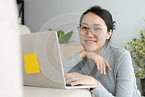 Asian woman is working on a laptop in the living room. She sit on the floor and sofa is office desk. On her face are smile and