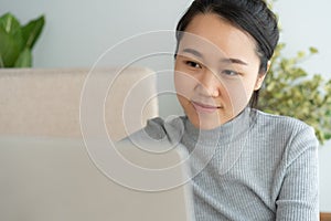 Asian woman is working on laptop in the living room. She sit on the floor and sofa is office desk. On her face are smile and