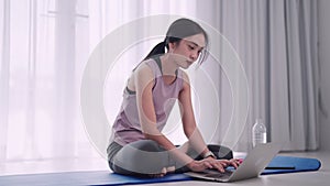 An Asian woman in workout attire uses her laptop to find exercise information on a yoga mat in her home