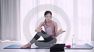 An Asian woman in workout attire uses a tablet for yoga lessons on a mat in her home's sitting room.