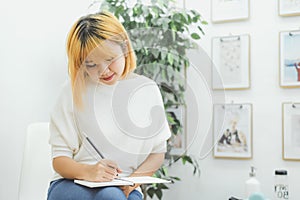 Asian woman write shopping lists in notepad by pen on her kitchen counter at home.