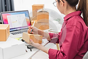 Asian woman writing and jotting down information From laptop to packaging box, online shopping, e-commerce and logistics concept