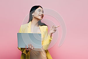 Asian woman in yellow outfit holding laptop and pointing with finger away  on pink