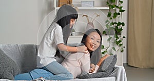 Asian woman young loving attentive mother lies on couch carries on her back little active jumping girl playful daughter