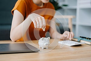 Asian women's hand saving a coin into piggy bank with wooden to calculateon and tablet on the table for business, finance,