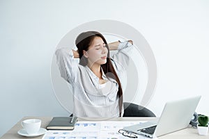 Asian women are tired from work on laptop sits in a chair stretching to relax and unwind while working hard at the office