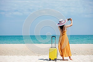 Asian women traveler holding yellow luggage walking and take a photo smart phone on the beach.