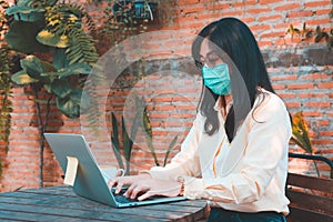 Asian women wearing face mask in Coronavirus COVID-19 pandemic works at laptop while sitting on bench in cafe.