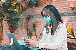 Asian women wearing face mask in Coronavirus COVID-19 pandemic works at Smartphone and laptop while sitting on bench in cafe.