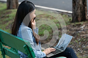 Asian working woman talking on smartphone and typing on laptop computer while sitting on bench in the public park