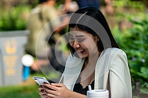 Asian working woman texting at smartphone while sitting on bench in the public park