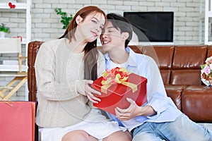 Asian young beautiful female girlfriend sitting smiling opening unboxing red present gift wrapped box on cozy sofa from handsome